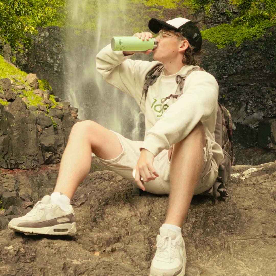 Man resting by a waterfall, drinking from a Jaje reusable bottle while hiking